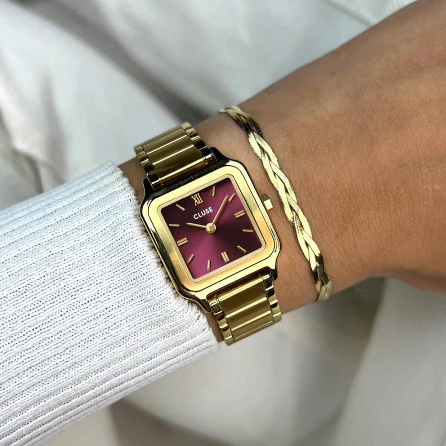 a gold coloured cluse watch with a  maroon face paired with a gold bracelet on girls wrist